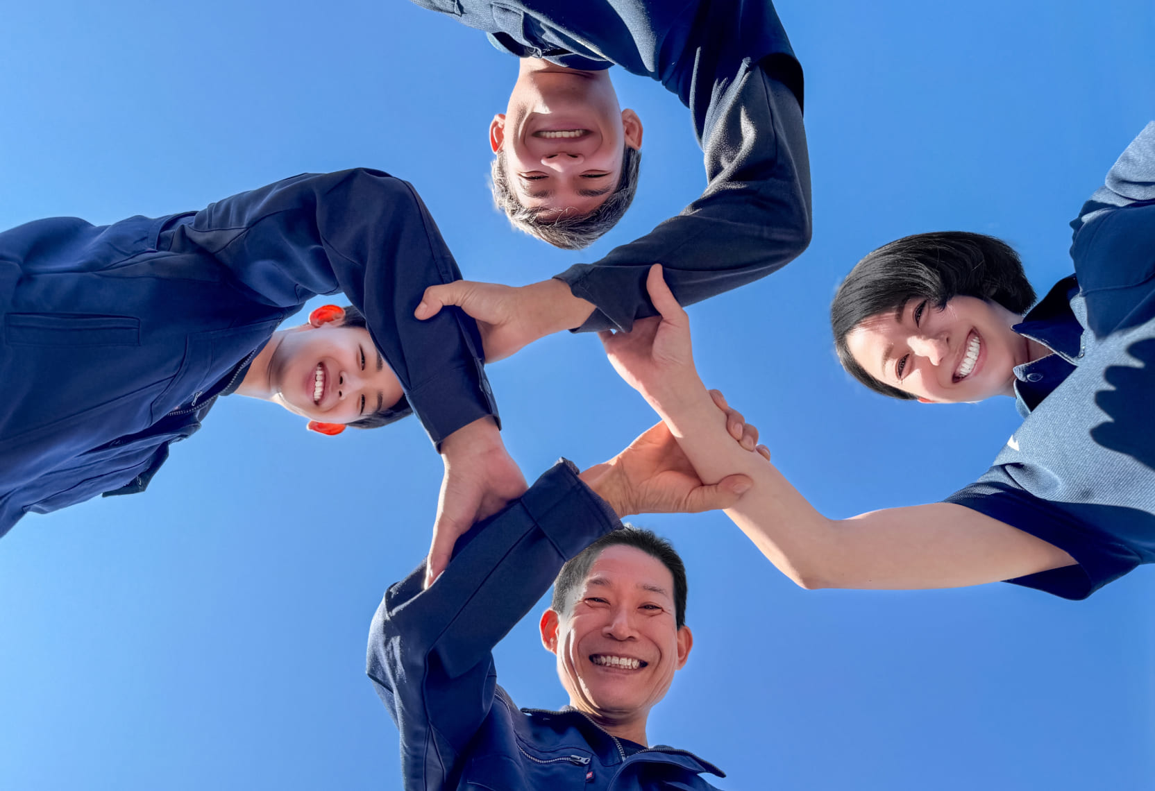 青空の下で4人の人々が手を組んで円を作っている様子が下から見上げた視点で写した画像。4人は青い作業着を着ている。