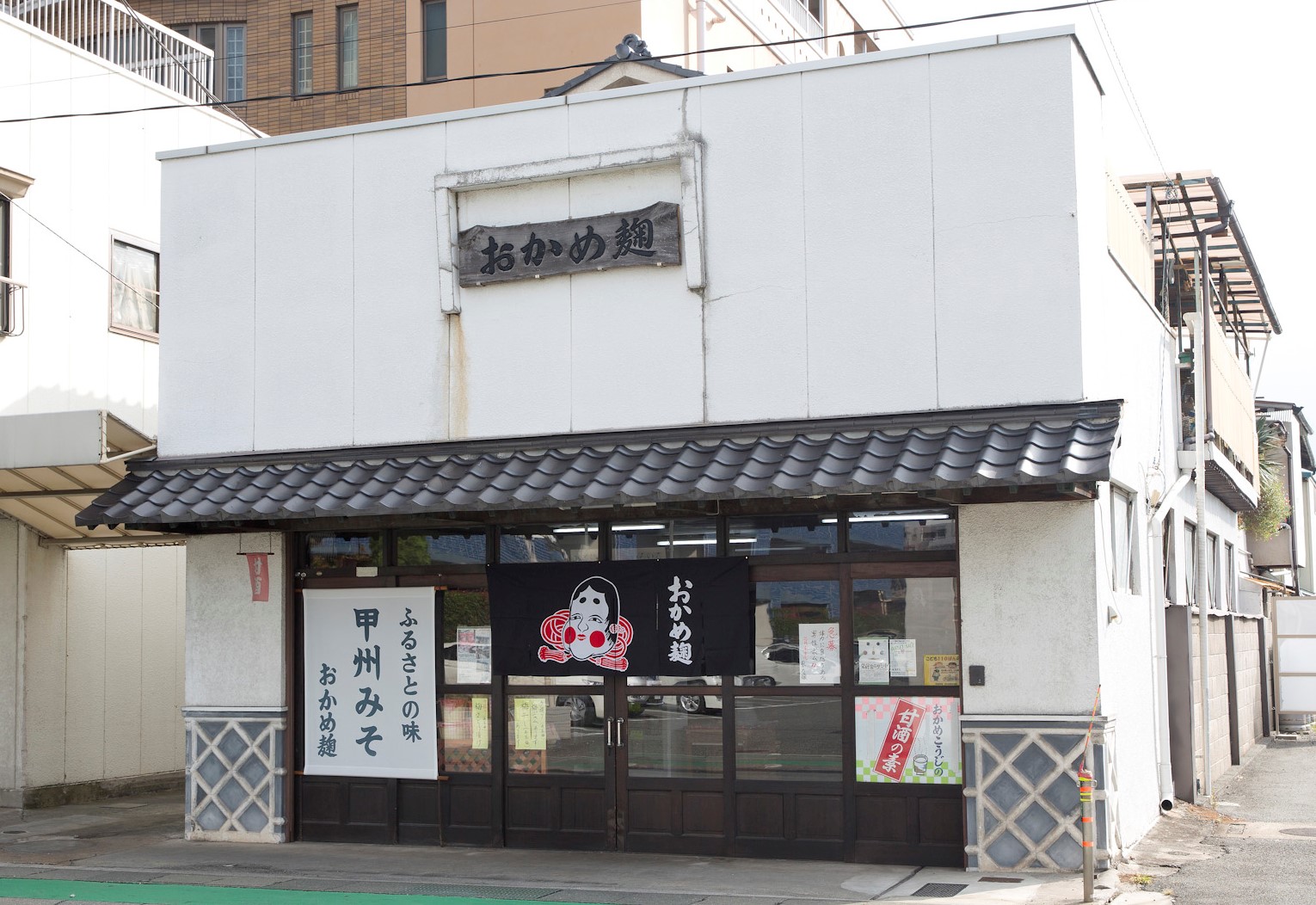 おかめ麹の看板とのれんが見える甲府市若松町の店舗外観