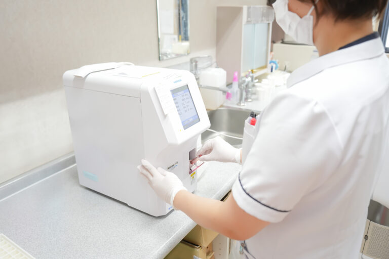 公式スタッフ紹介に掲載された検査室の機器と後ろ姿の職員